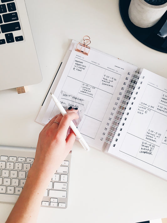 flatlay image of a woman's hand holding a white pen and writing in her planner white keyboard laptop